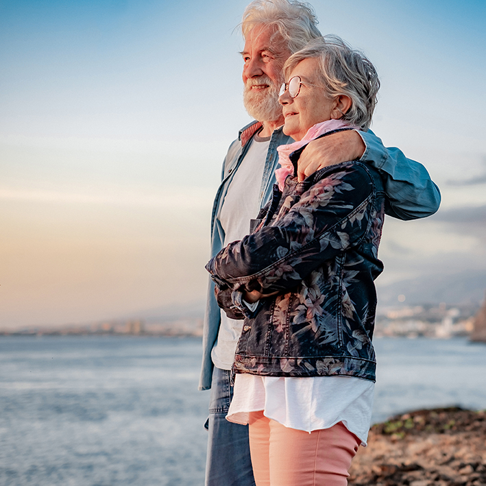 due pensionati residenti all’estero che si abbracciano sulla spiaggia guardando il mare